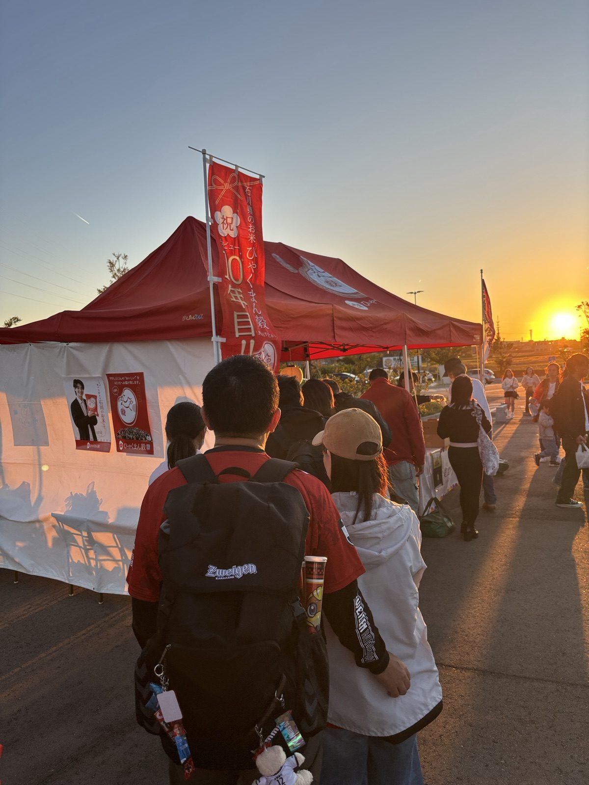 夕日に照らされるJAのおにぎり配布と五郎島金時の大学芋試食のテントに並ぶサポーター